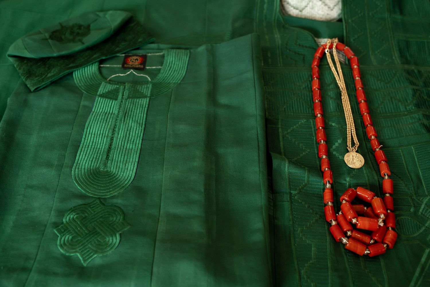 Green embroidered African fabric with red coral beaded necklace and gold coin pendant displayed elegantly.