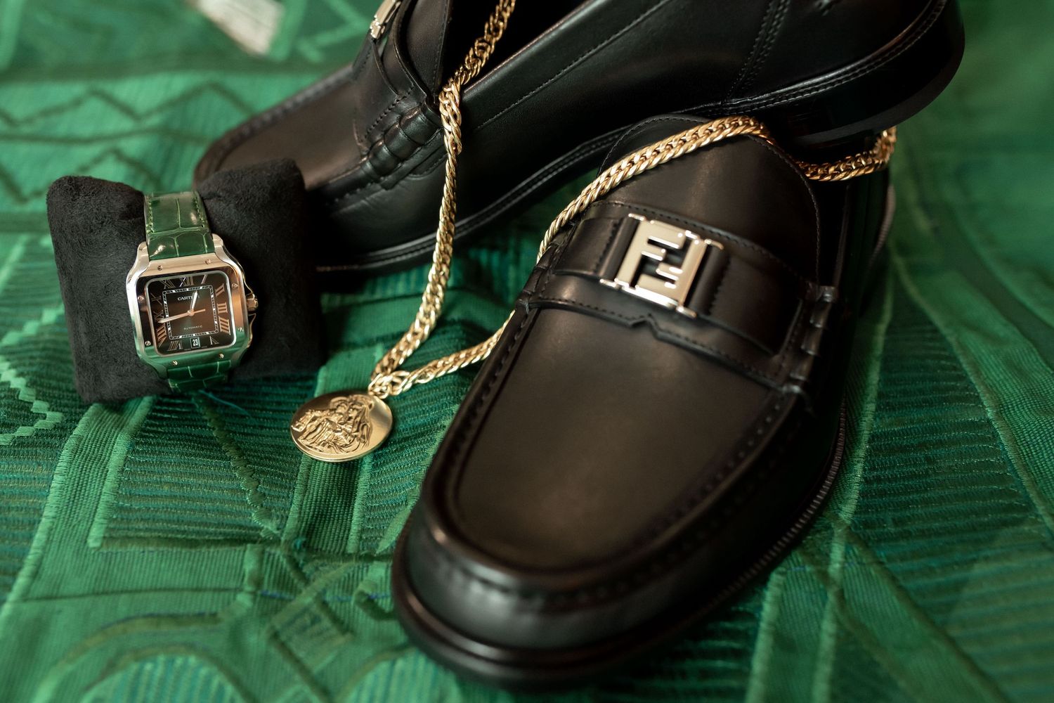 Black Fendi loafers with gold FF logo beside a gold chain necklace and luxury watch on green fabric.