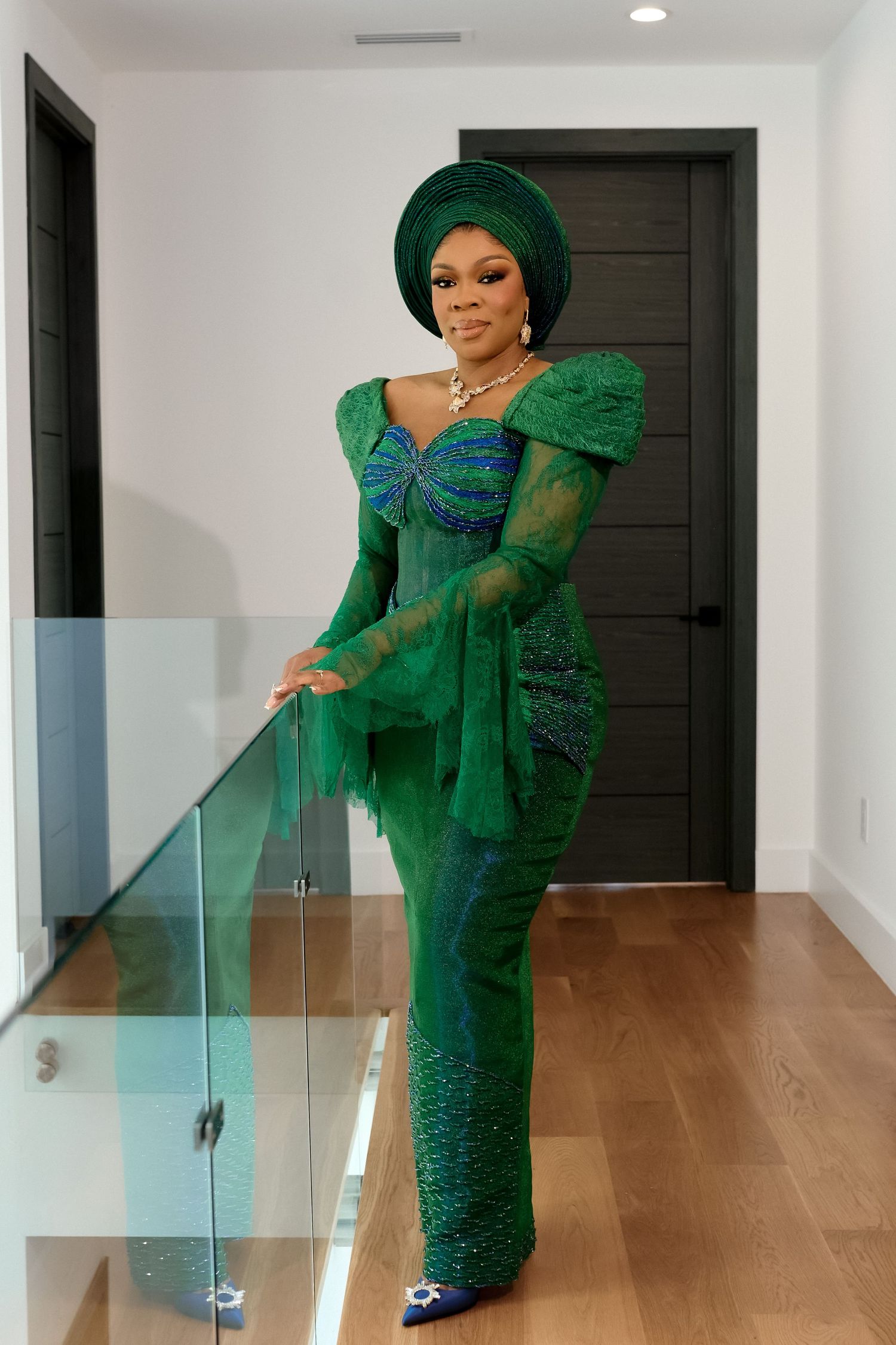 Woman in elegant green Nigerian traditional attire with gele headwrap poses gracefully indoors.