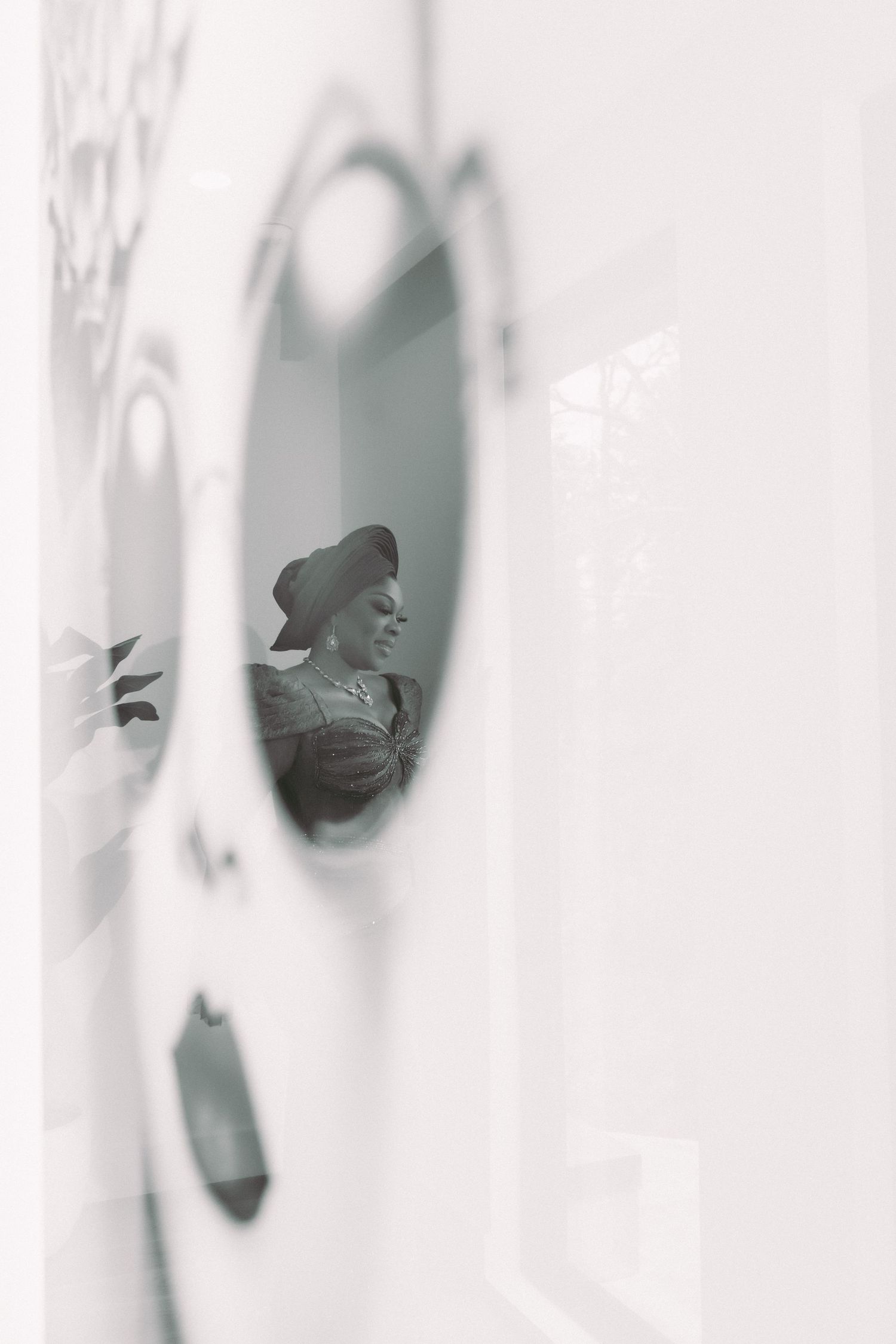 Black and white photo of a woman reflected in a round mirror, wearing a head wrap and elegant attire.