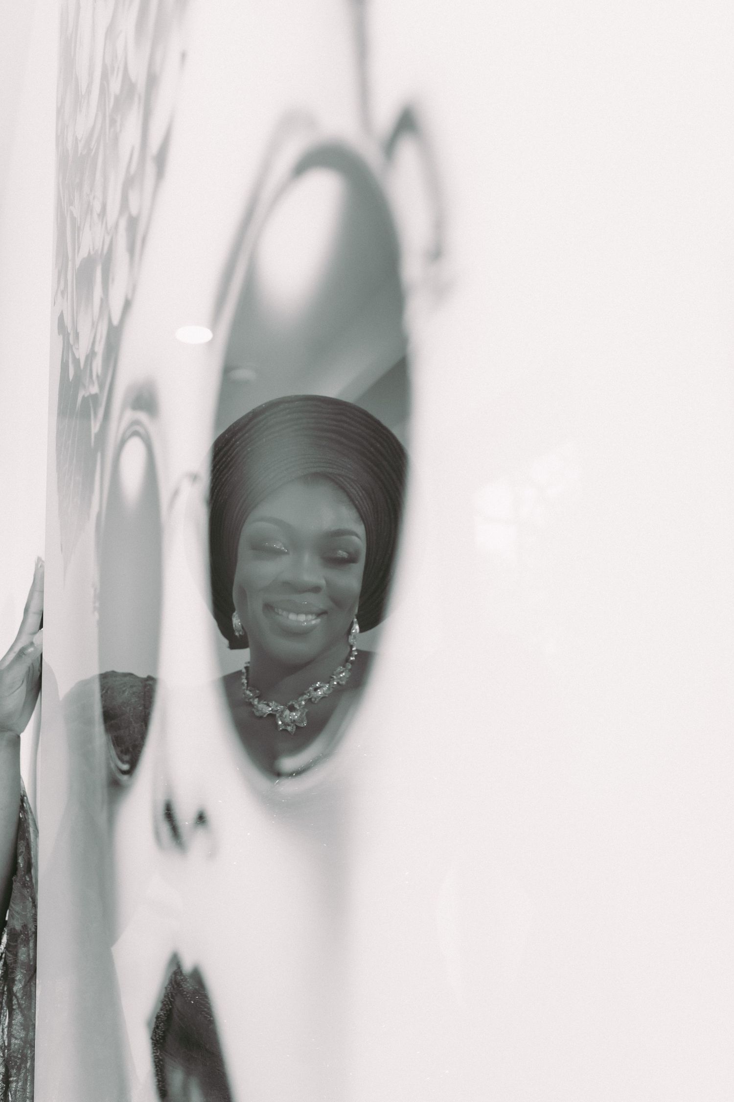 Black and white photo of a smiling woman reflected in a round mirror, wearing a necklace and elegant headpiece.