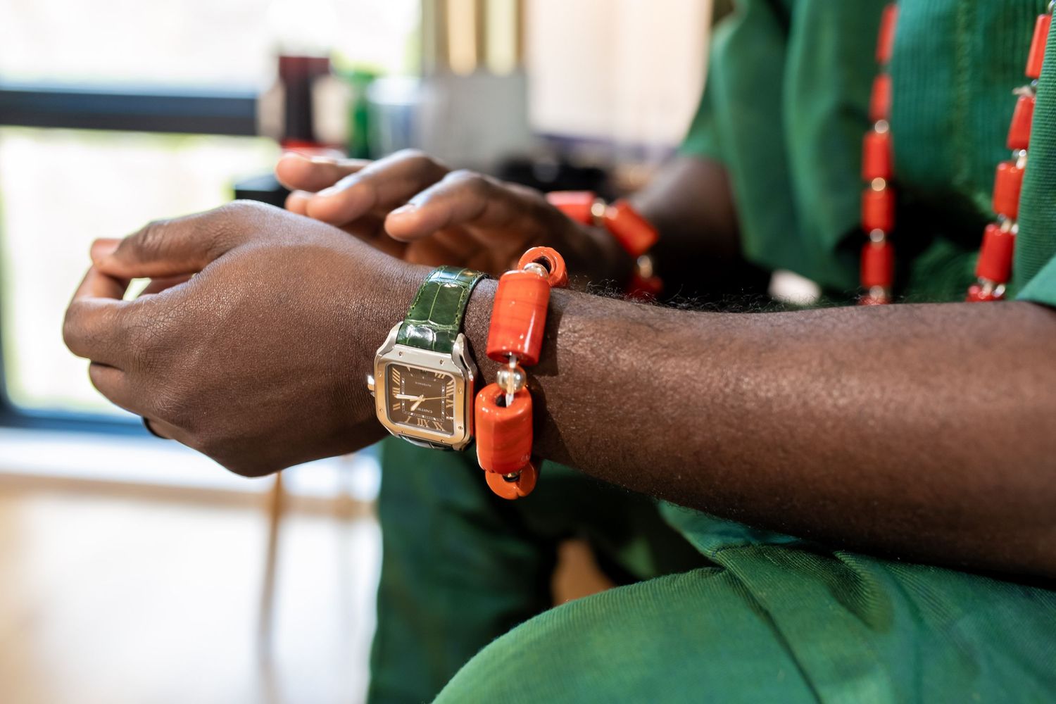 Close-up of a person wearing a square-faced watch and orange beaded bracelet on their wrist, dressed in green clothing.