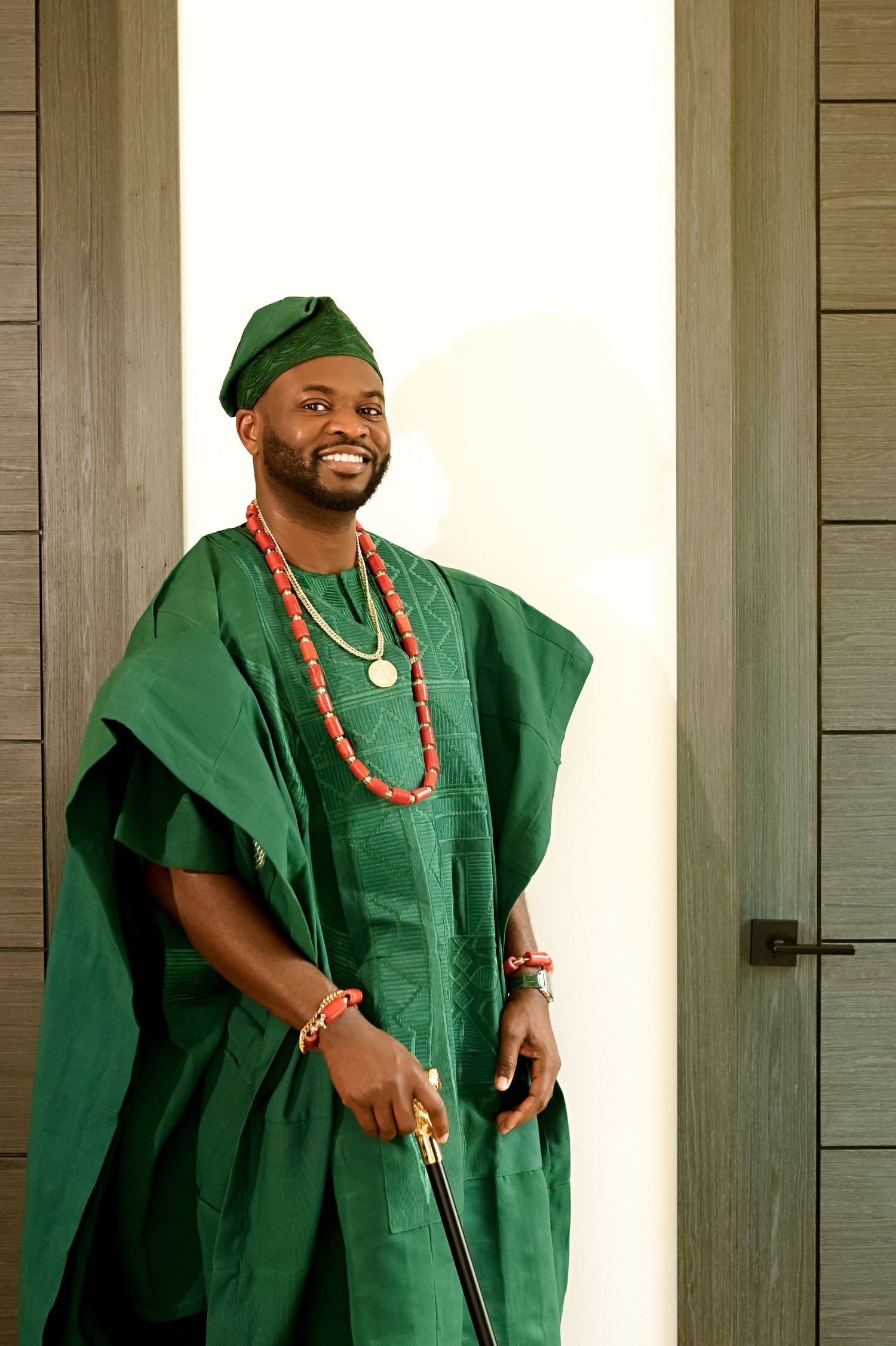 Man smiling in green traditional African agbada outfit with red coral beads necklace and matching cap.