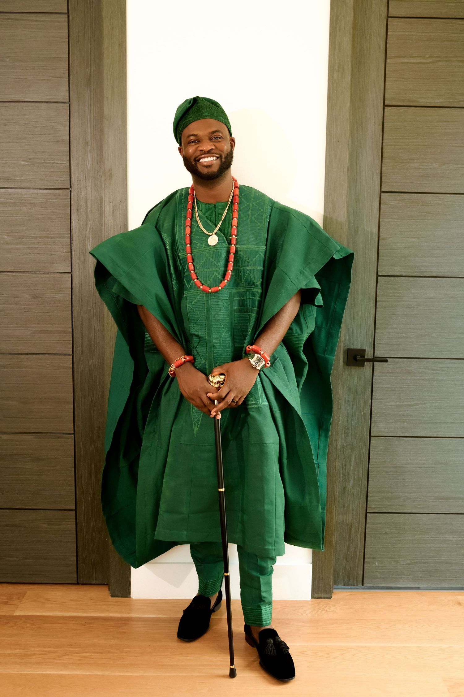 Man smiling in green traditional African Agbada outfit with red beaded necklace, matching cap, and cane.