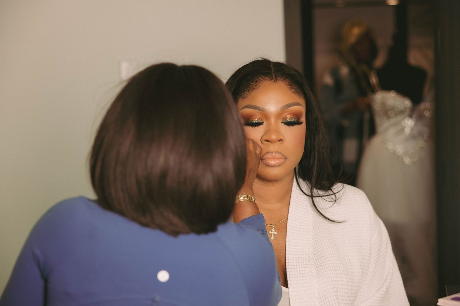 Bride getting makeup applied by a makeup artist while wearing a white robe, with a wedding dress visible in the background.