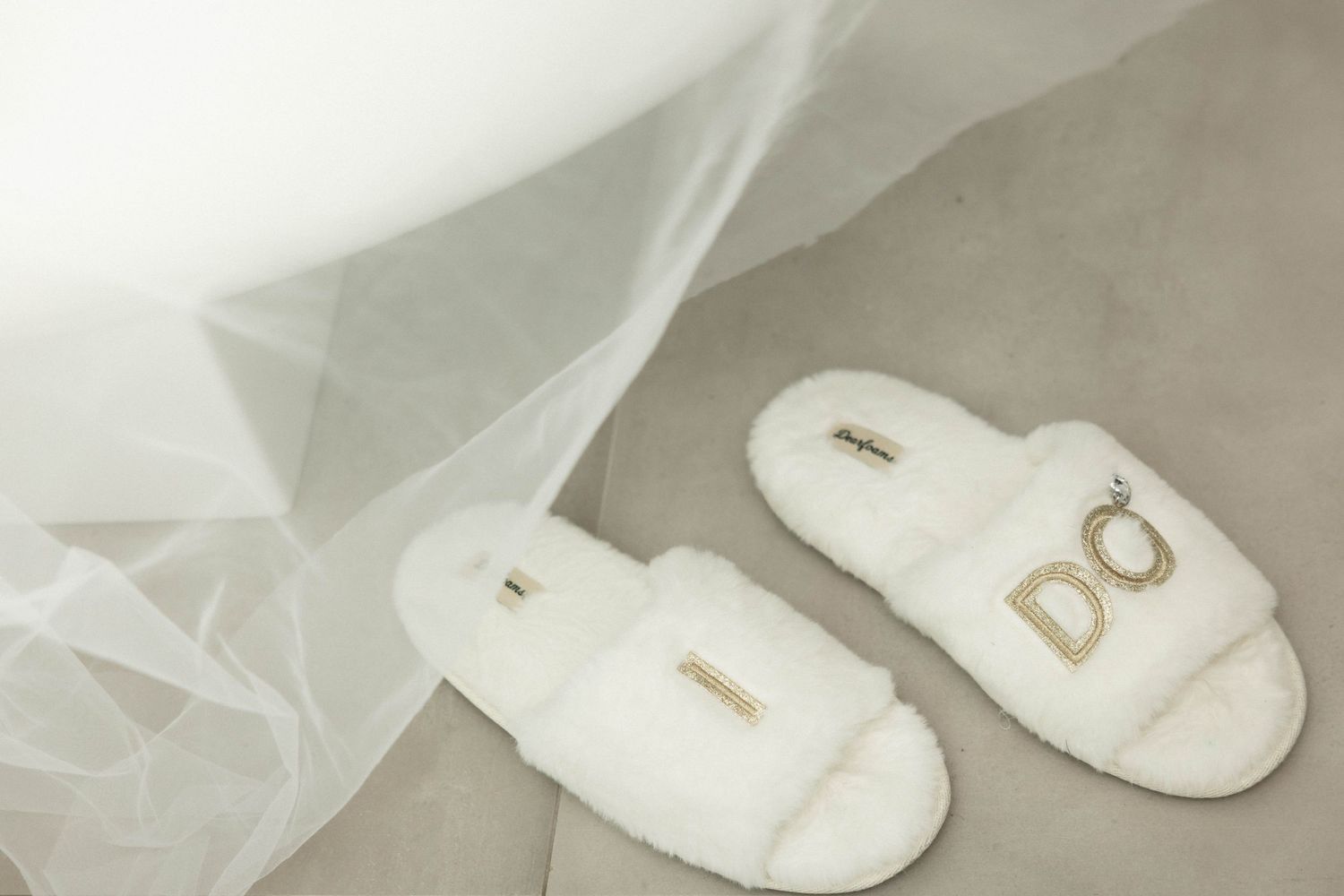 White fluffy bridal slippers with gold DO letters on gray floor next to wedding veil.
