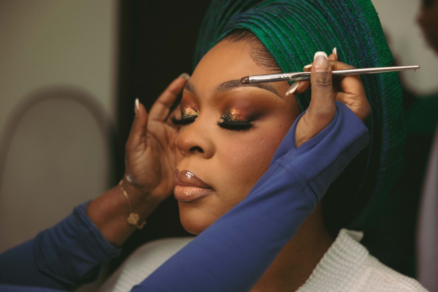 A makeup artist applies eyeshadow with a brush on a woman wearing a green head wrap and glamorous eye makeup.
