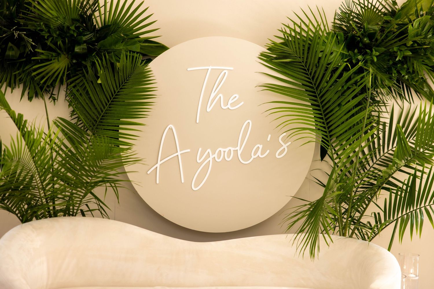 Elegant circular sign reading The Ayoola's surrounded by lush tropical palm leaves on a neutral background.
