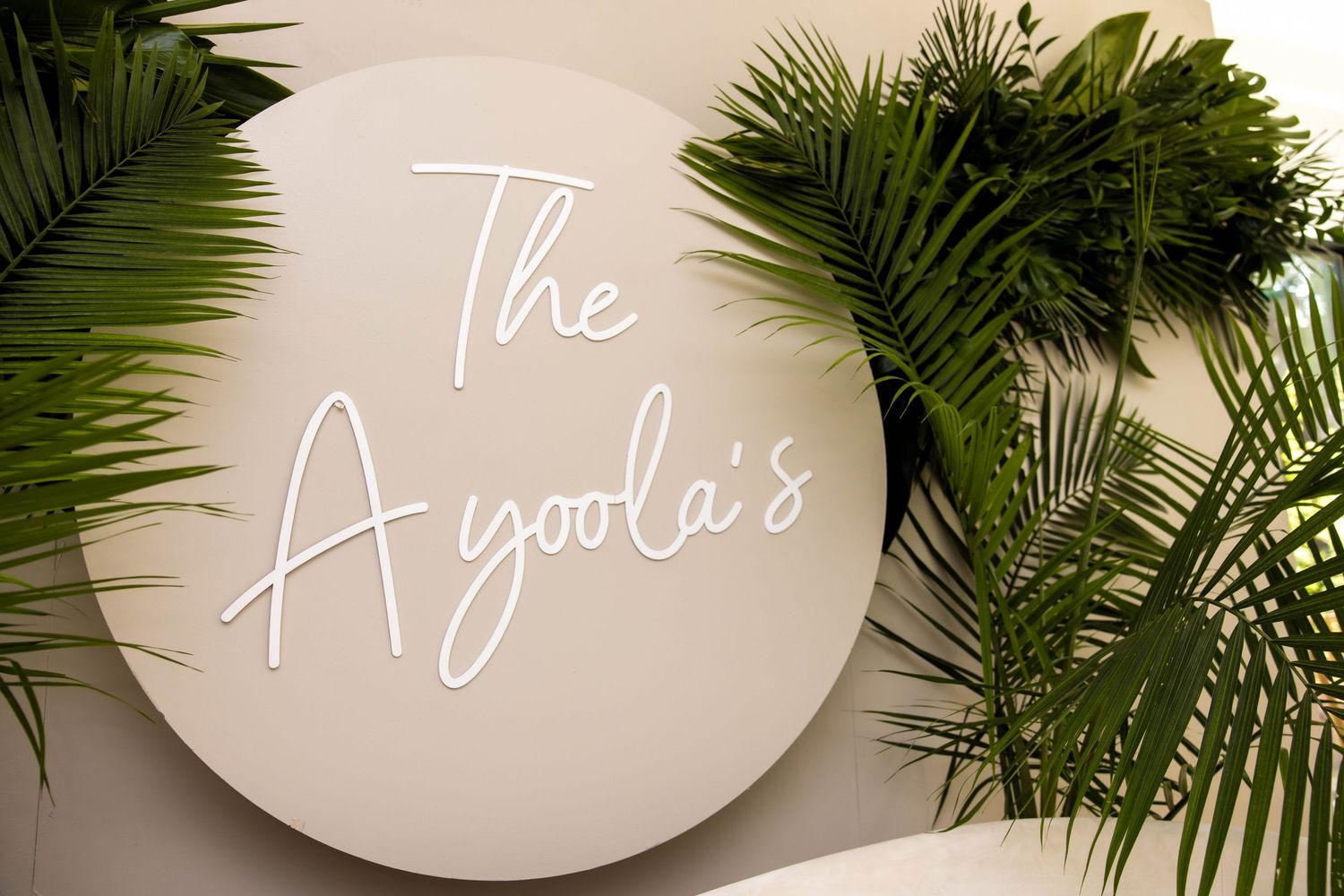 Round white sign reading The Ayoola's in neon script, surrounded by tropical palm leaves on a light wall.
