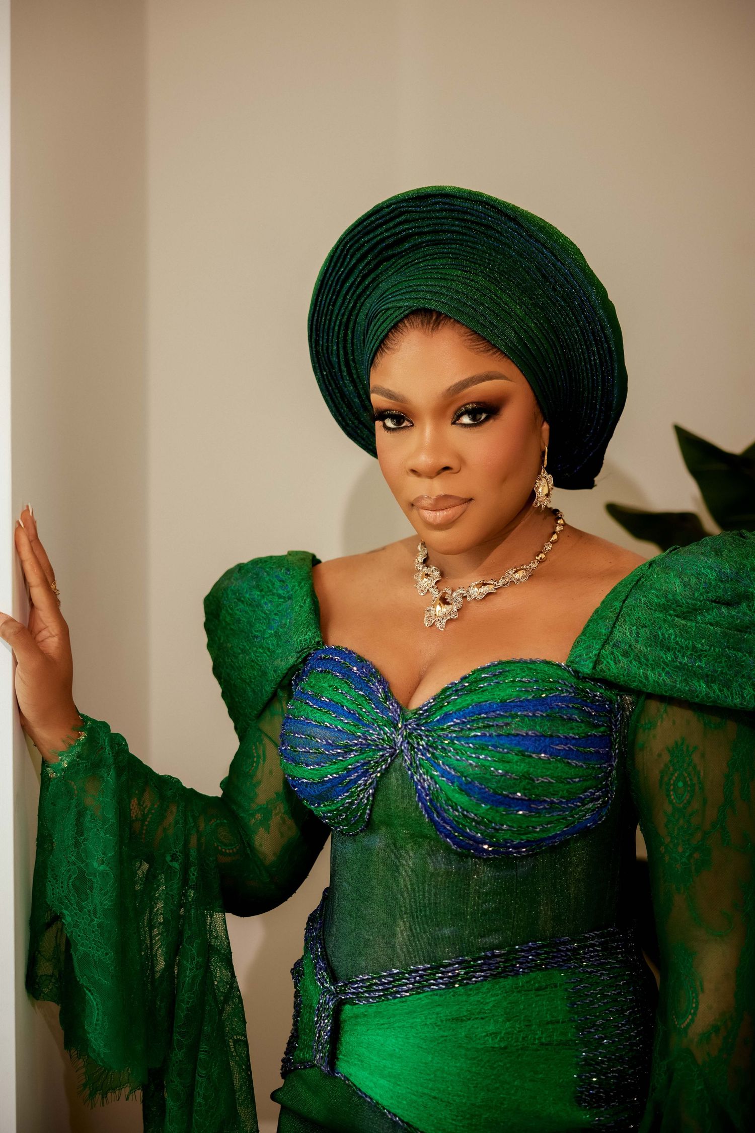 Woman in elegant green Nigerian traditional attire with gele headwrap and beaded bodice poses gracefully.