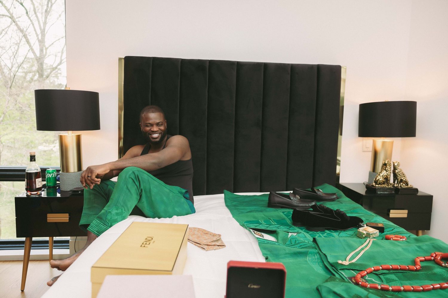 Smiling man sitting on a bed with green fabric, jewelry, and luxury items displayed in an elegant bedroom.