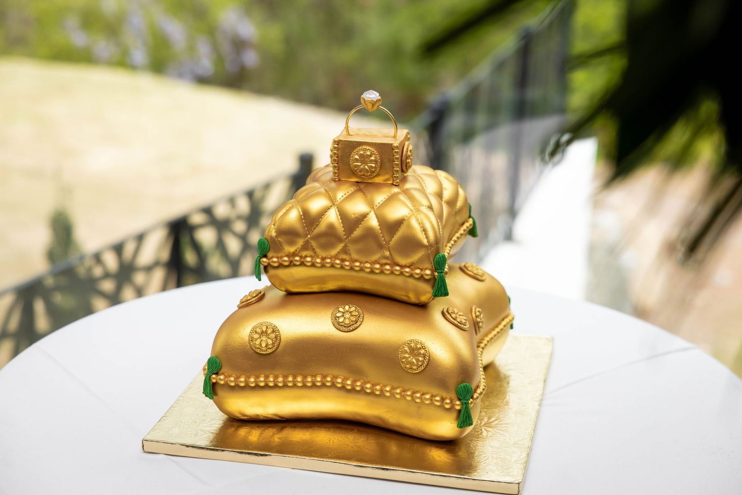 Three-tier golden quilted pillow wedding cake with emerald green accents and a ring topper on a white table.