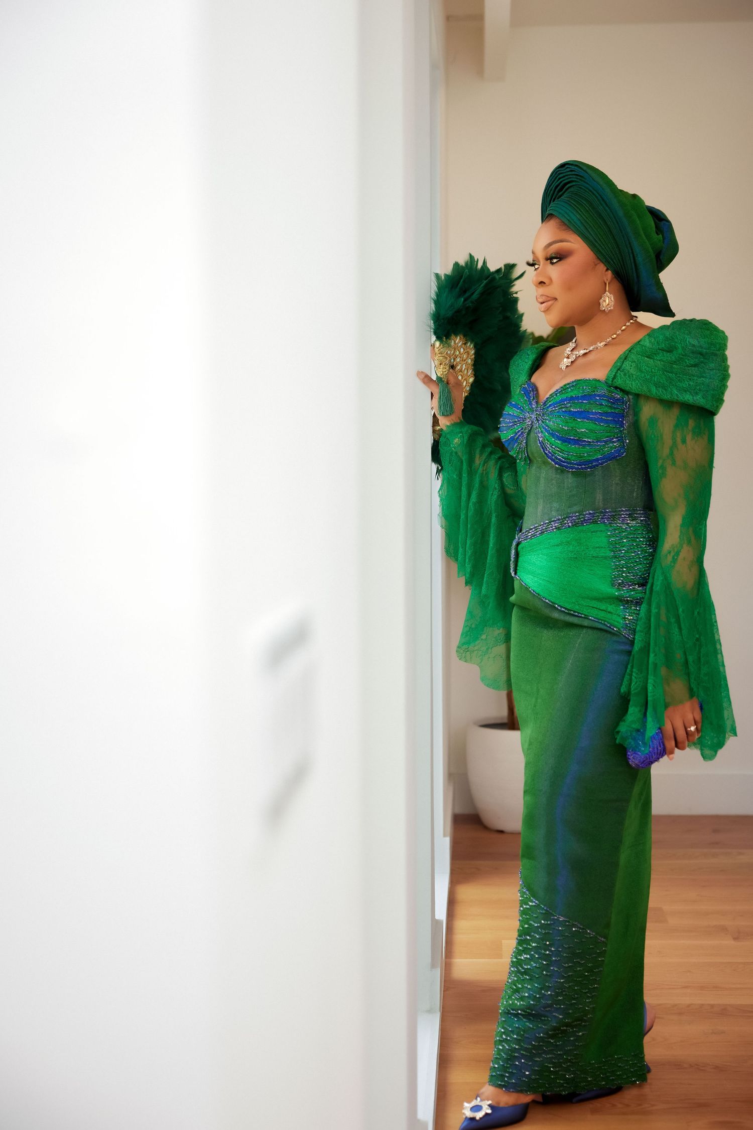 Woman in elegant green Nigerian traditional attire with gele headwrap and feather fan posing in doorway.