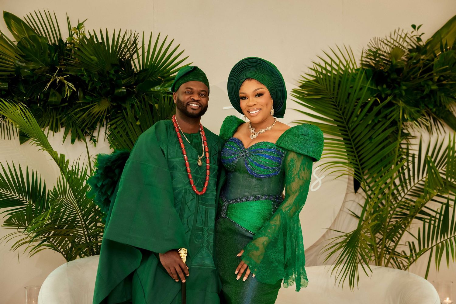Couple in matching green traditional Nigerian attire with gele headwrap and agbada, posing before tropical foliage backdrop.