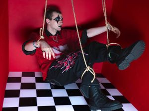 Person in black mask and red shirt suspended by ropes in a red room with checkered floor.