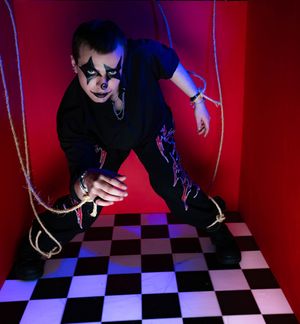 Child in clown makeup and black costume poses dramatically in red box with checkered floor and rope strings.
