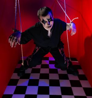 A person in creepy clown makeup and black outfit posed as a marionette puppet with strings on a checkered floor.