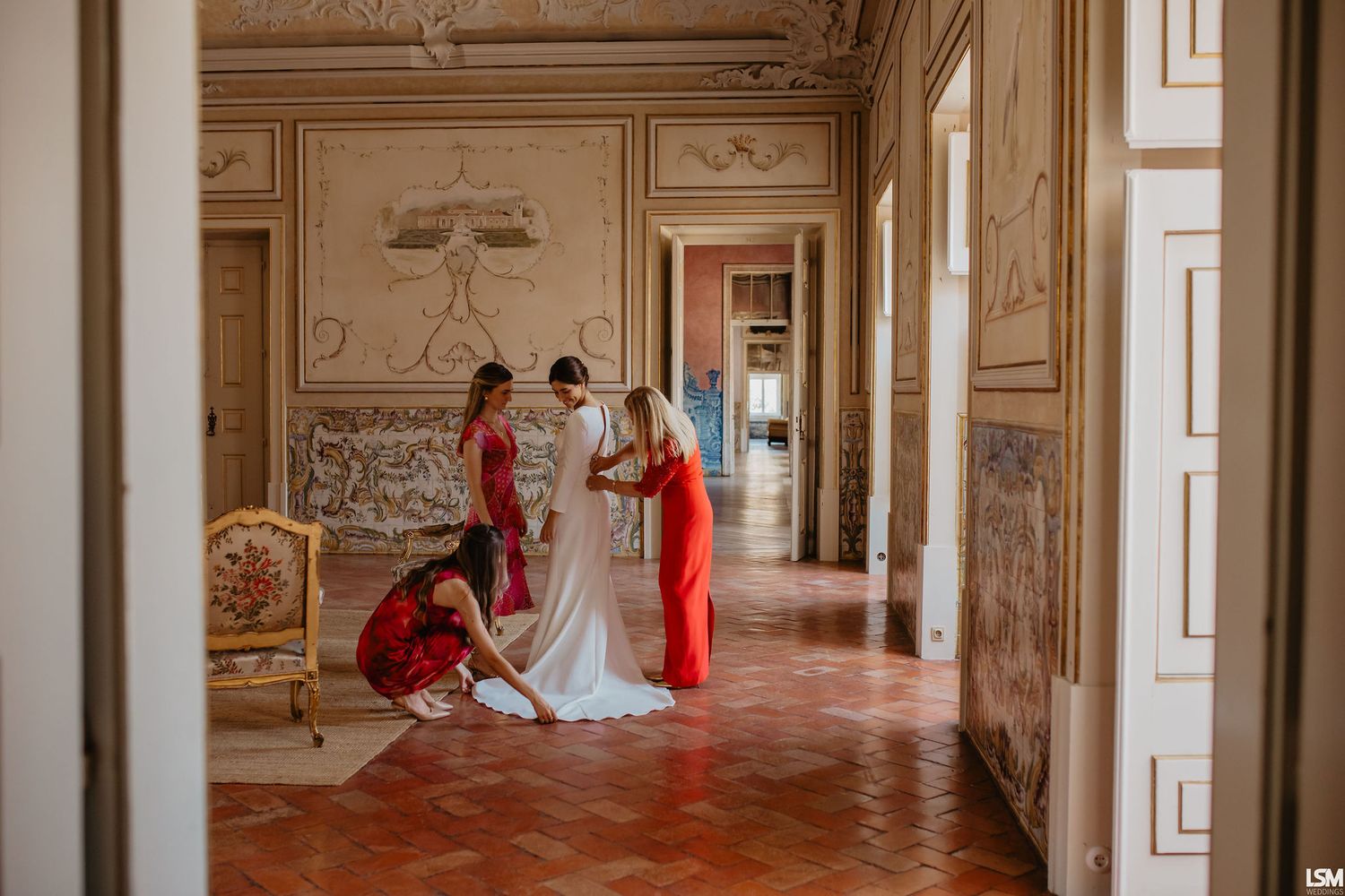 Bridesmaids in red dresses help bride prepare in elegant ornate hall with frescoed walls and terracotta floors.