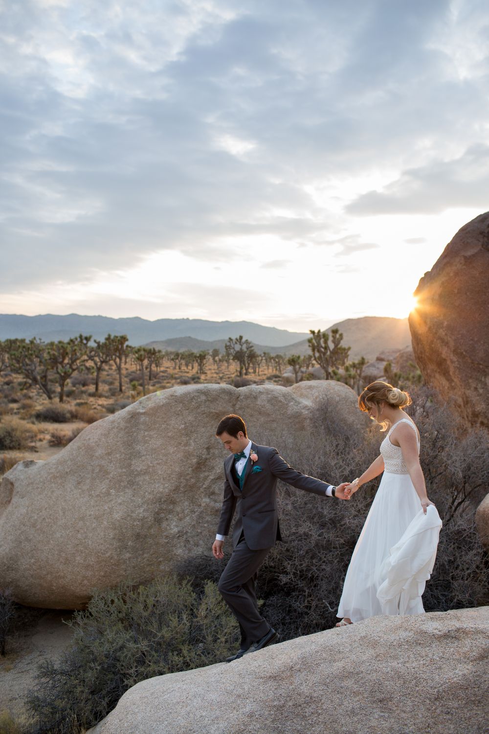 A young couple climbs down a boulder in Joshua Tree National Park in wedding attire.