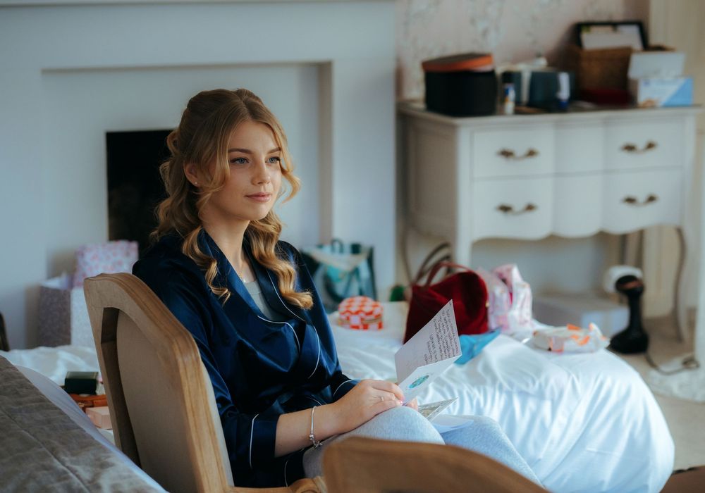 Young woman in blue velvet outfit sits thoughtfully holding papers in an elegantly decorated bedroom.