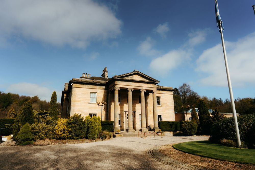 Grand neoclassical stone mansion with Greek columns, surrounded by manicured gardens and trees under a blue sky.