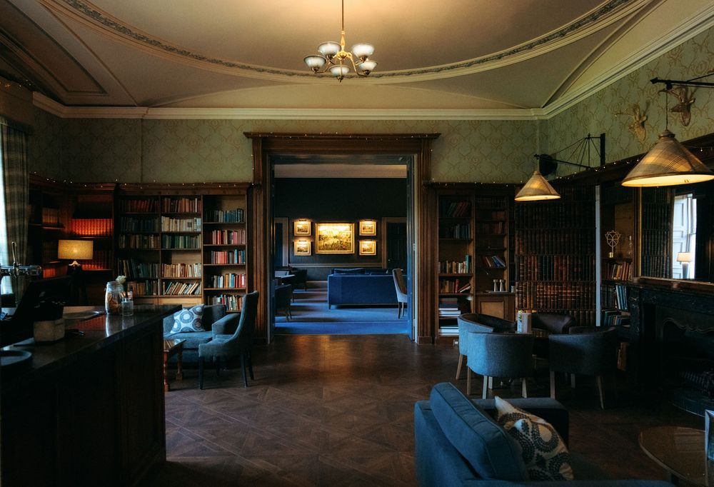 Elegant library room with floor-to-ceiling bookshelves, leather chairs, chandelier lighting, and a doorway leading to an art-filled hallway.