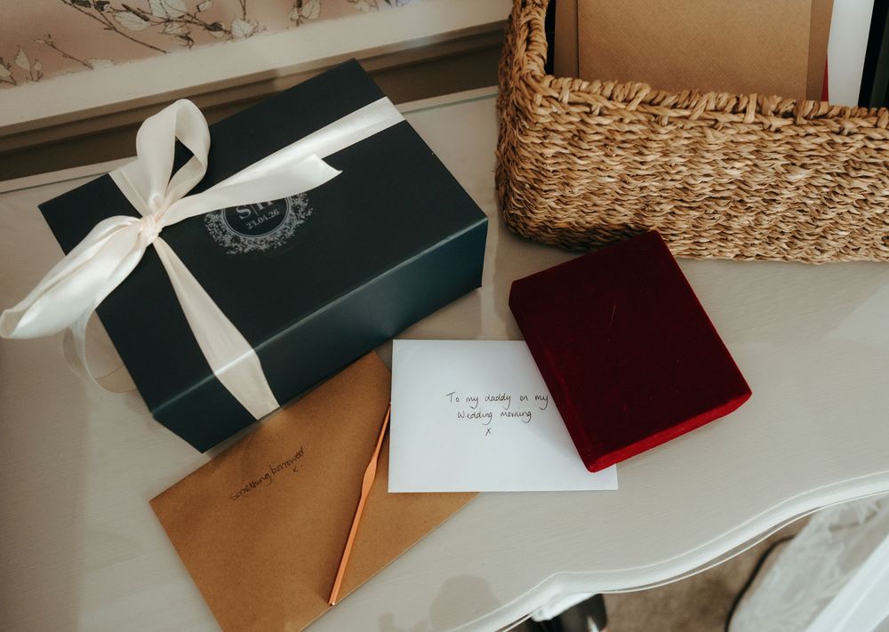 Elegant green gift box with white ribbon bow, kraft envelope, red velvet jewelry box, and wicker basket on white table.