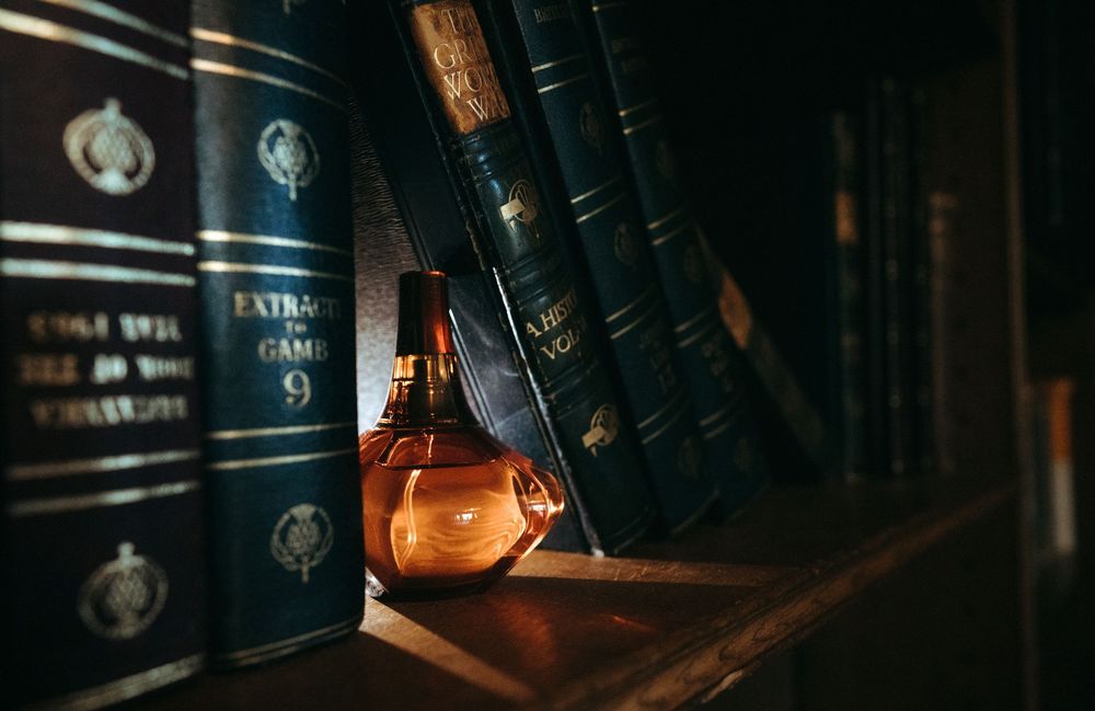 Amber glass perfume bottle leaning against vintage leather-bound books on a dark wooden shelf.