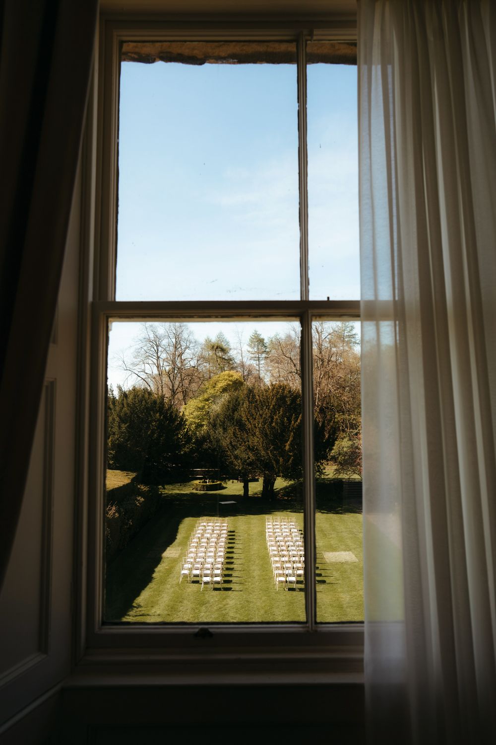 View through a tall window of an outdoor wedding ceremony setup with white chairs on a green lawn surrounded by trees.