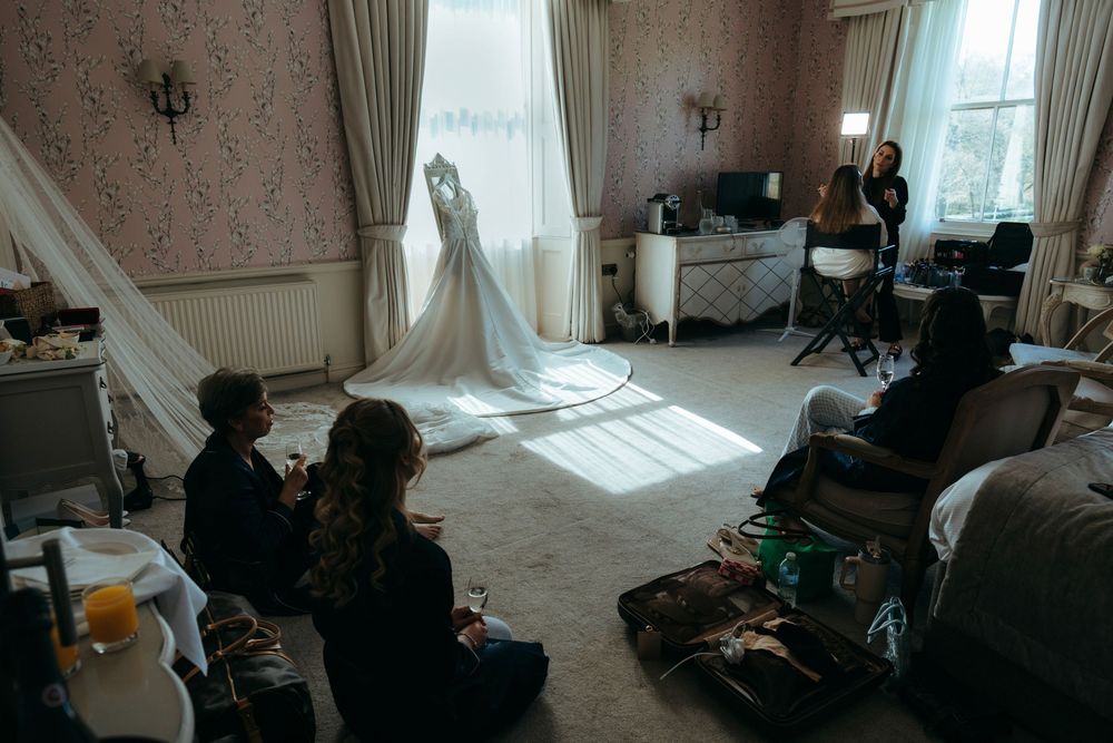 Bridal suite preparation with wedding dress displayed by window, bridesmaids seated on floor surrounded by luggage.