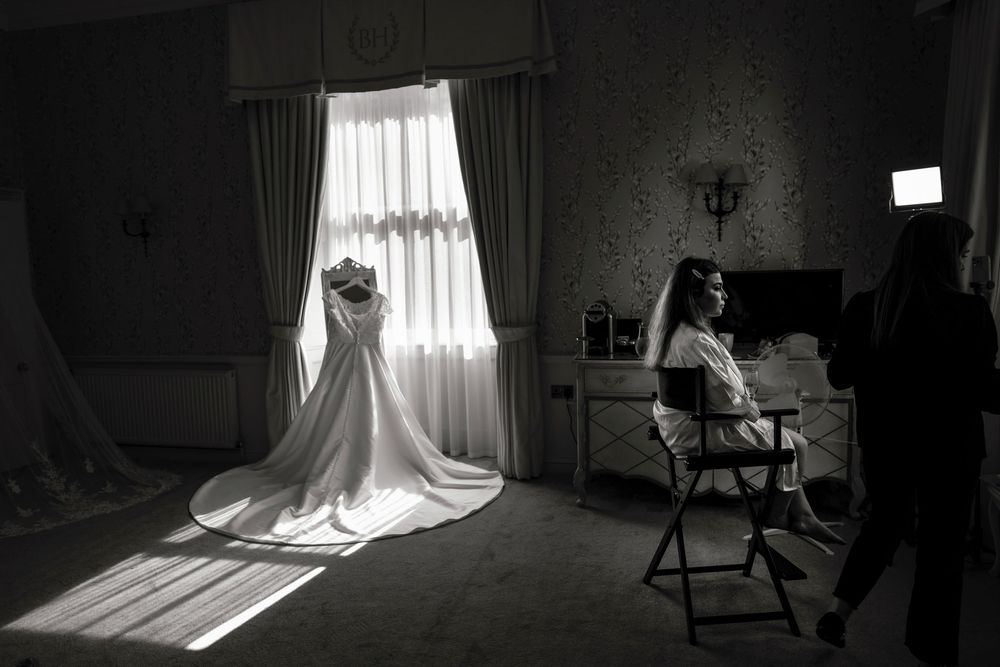 Black and white photo of a bride sitting in a chair beside a glowing wedding dress displayed near a sunlit window.