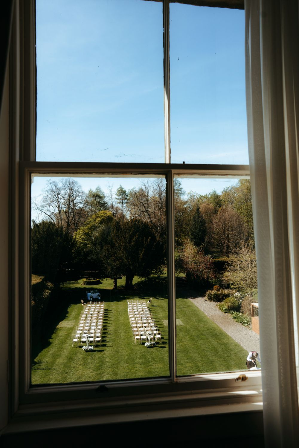 View through a window overlooking an outdoor wedding ceremony setup with white chairs on a green lawn surrounded by trees.