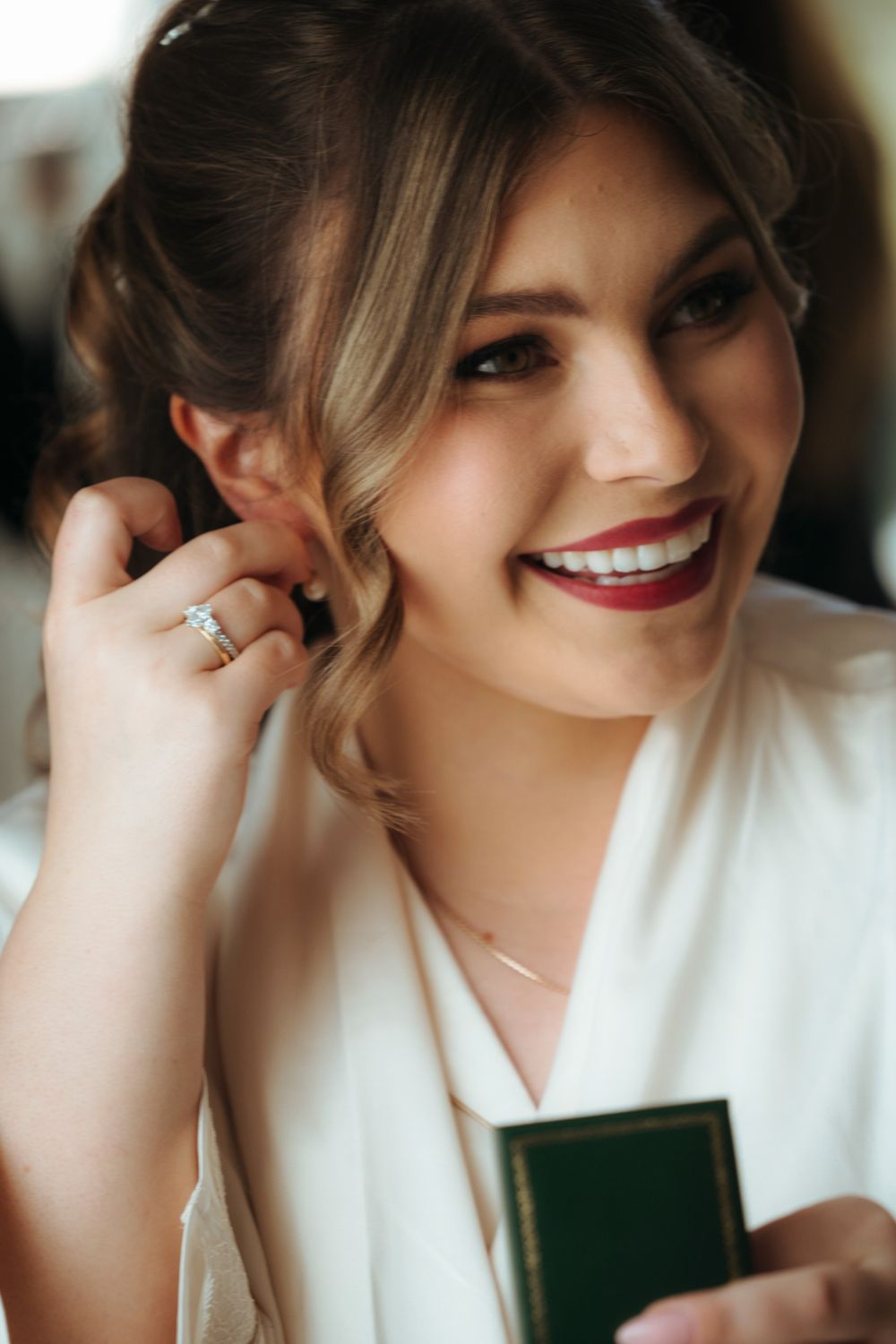 Smiling bride in white robe putting on earring, wearing engagement ring and delicate necklace, with elegant updo and red lips.