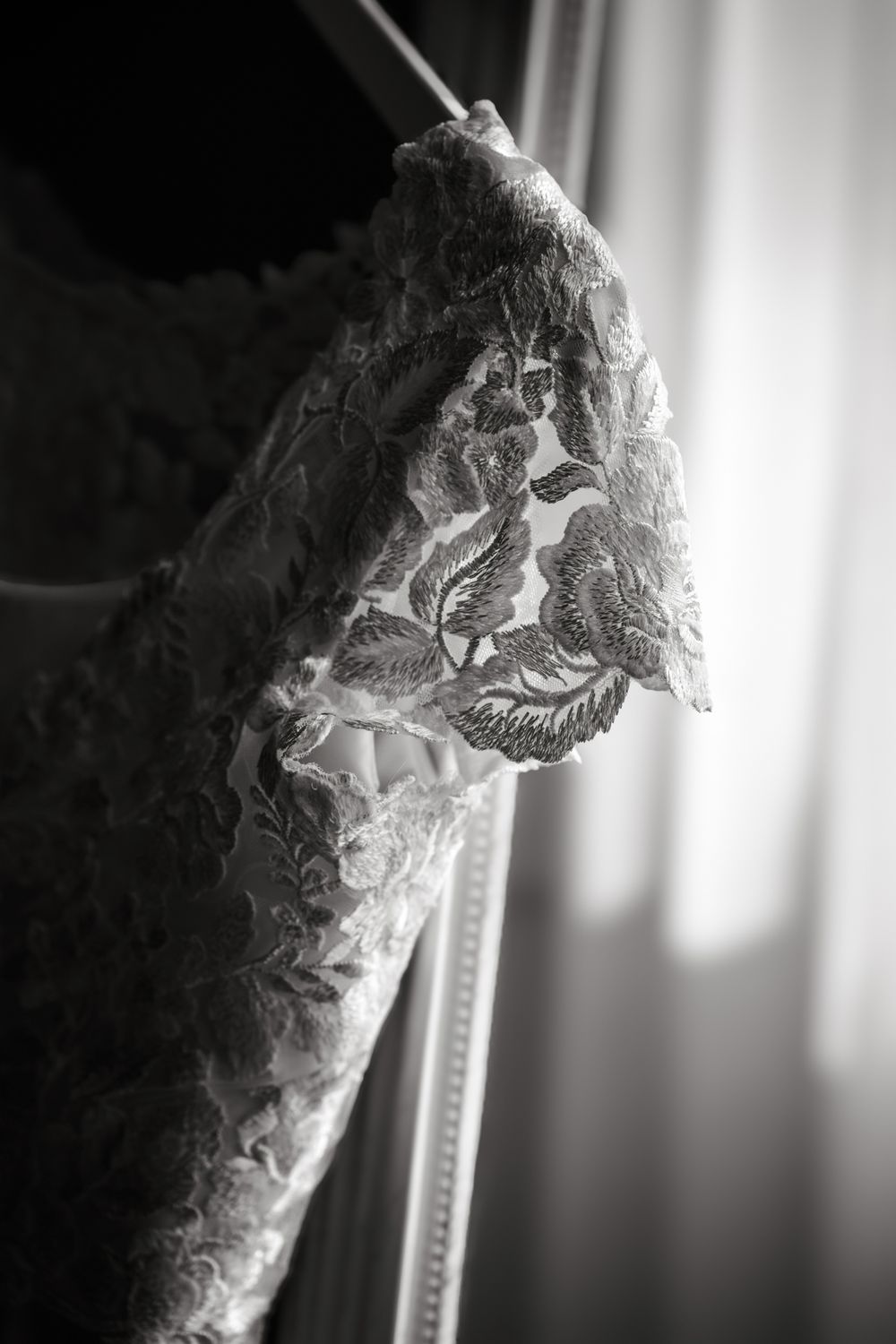 Black and white close-up of an elegant lace wedding dress sleeve with floral embroidery near a window.