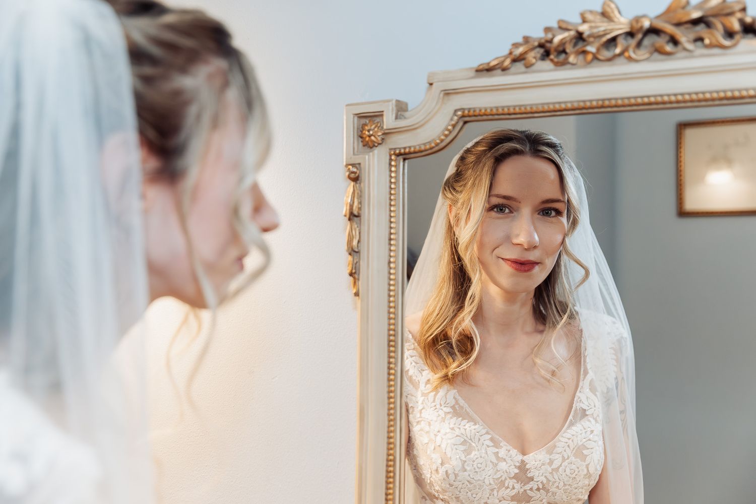 Bride in lace wedding dress and veil admiring her reflection in an ornate gold-framed mirror.