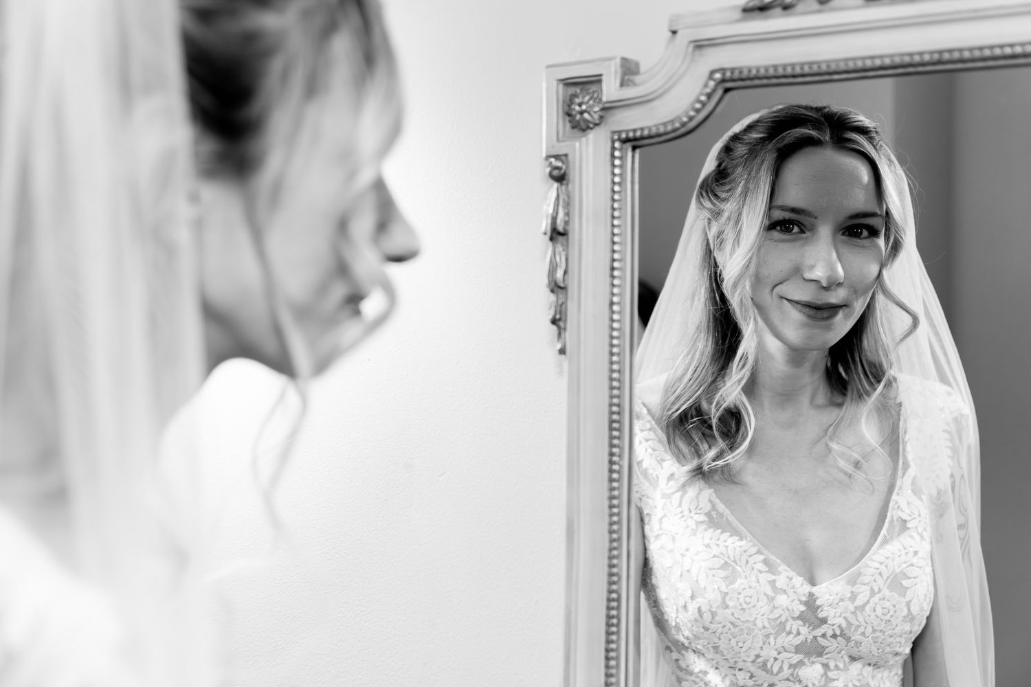 Black and white photo of a bride in lace gown admiring her reflection in an ornate mirror on her wedding day.