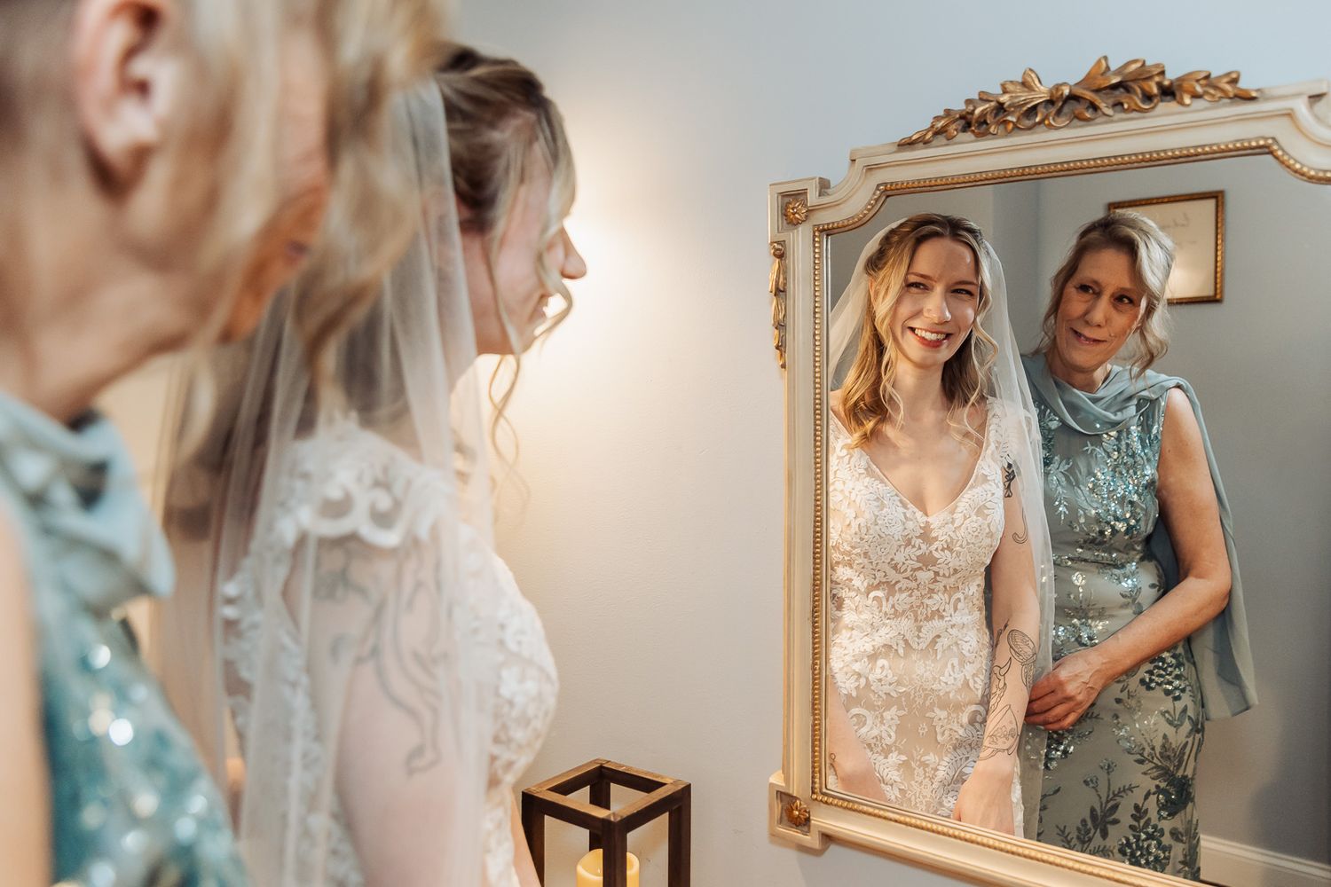 Bride in lace wedding dress smiles at reflection in ornate gold mirror while woman in blue dress stands beside her.