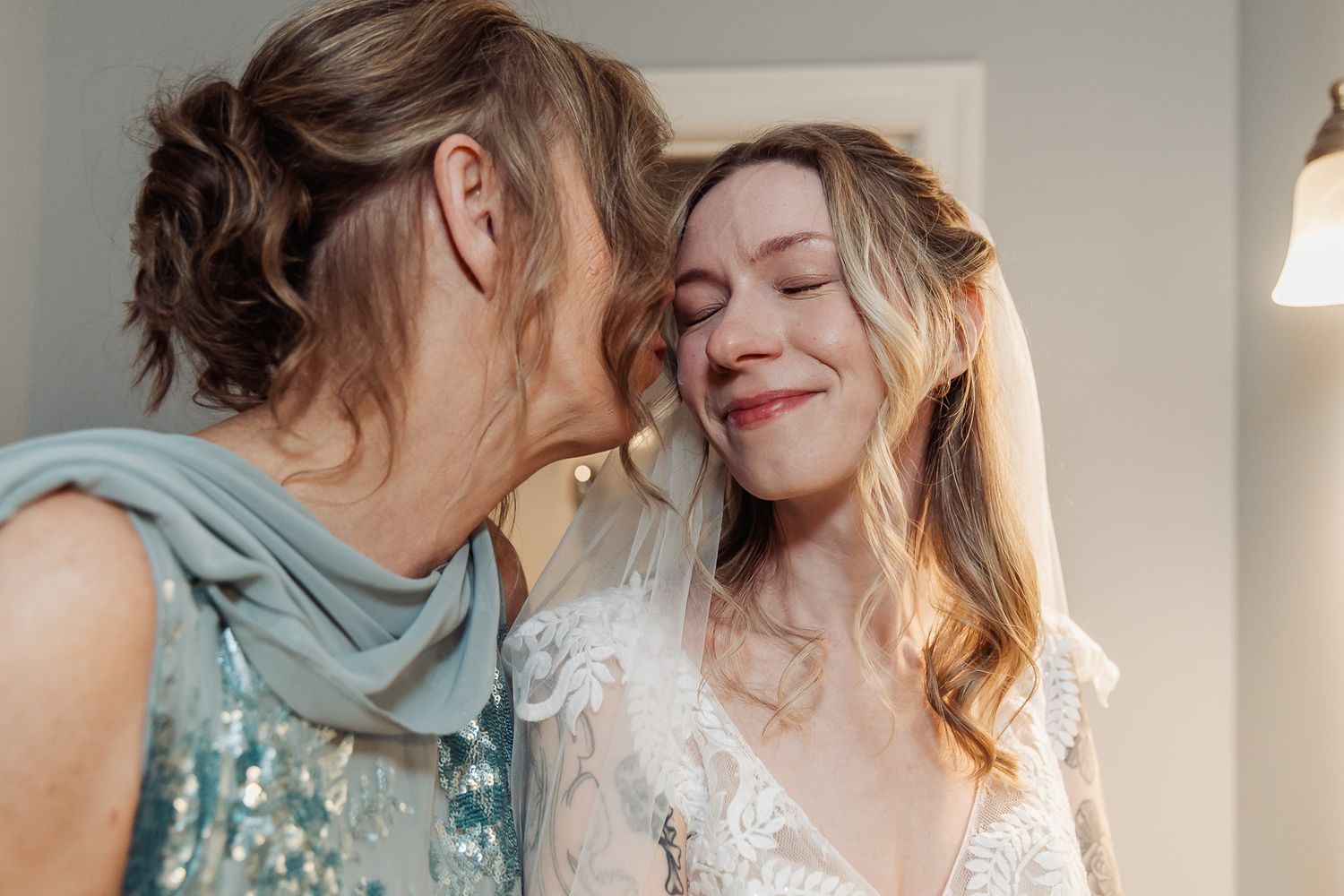 Mother kissing bride on cheek in tender wedding preparation moment, bride in lace gown smiling with eyes closed.