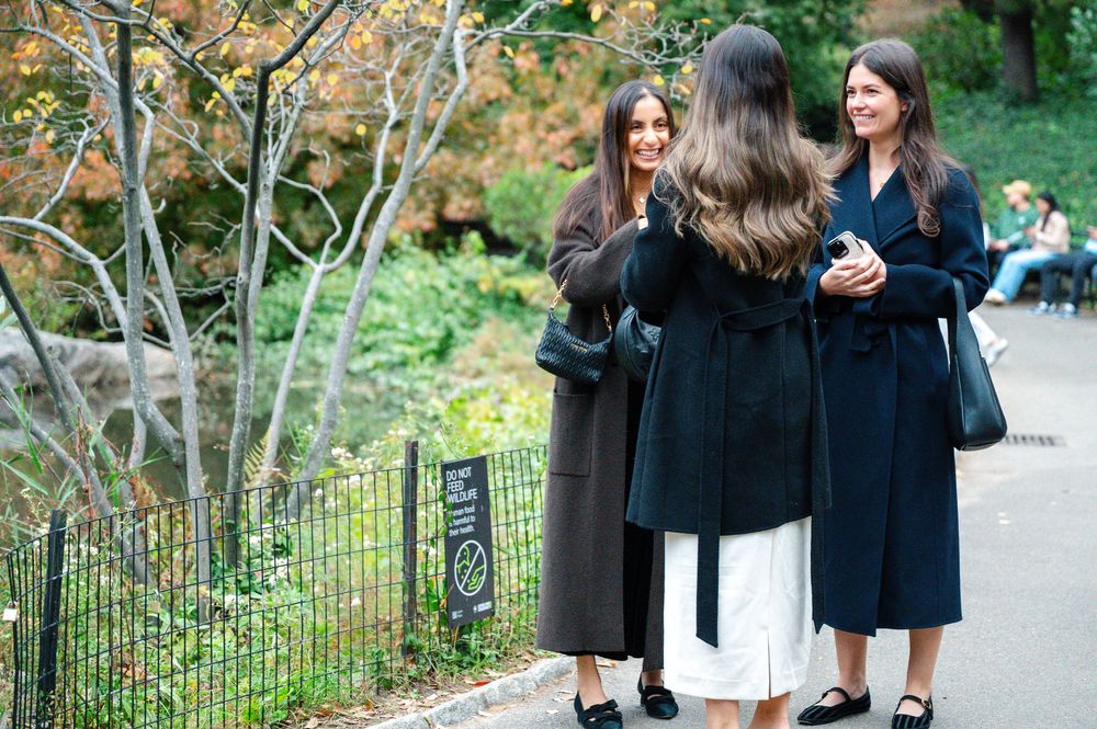 Three women in black coats chat on a park path surrounded by autumn foliage.
