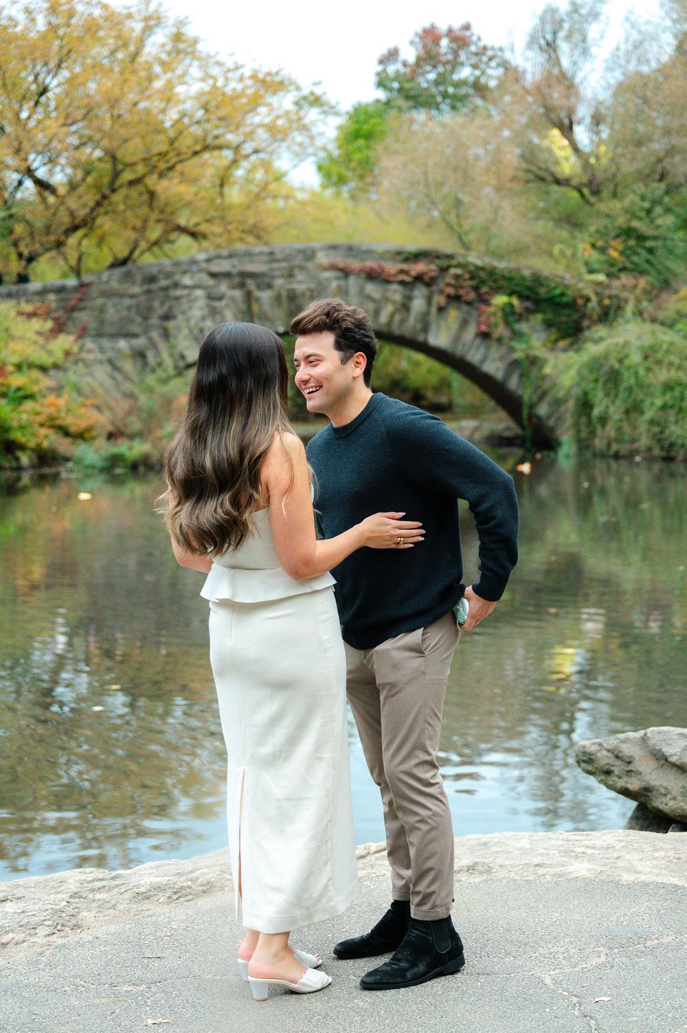 Couple laughing together by a stone arch bridge over a pond in Central Park during autumn.