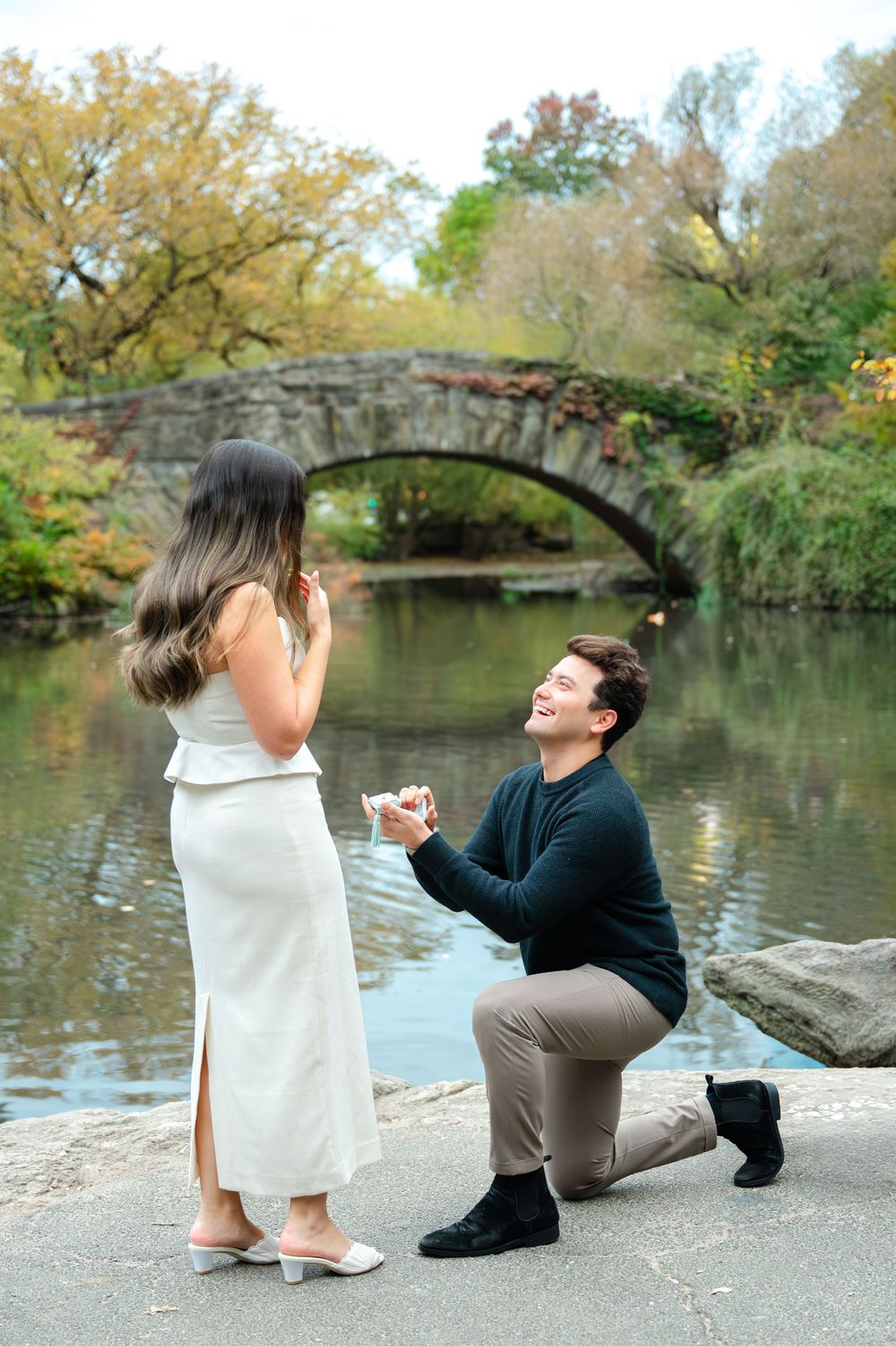 Man proposing on one knee with ring box by Central Park pond with stone arch bridge in autumn.