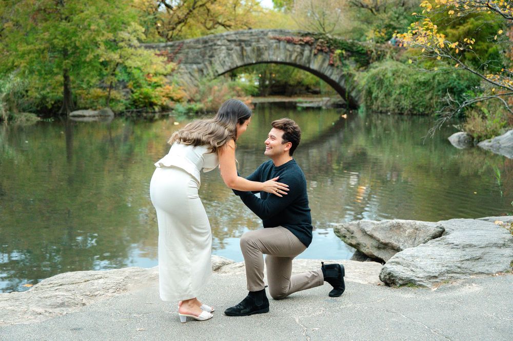 Man proposing on one knee to woman by a reflective pond with stone arch bridge in Central Park.