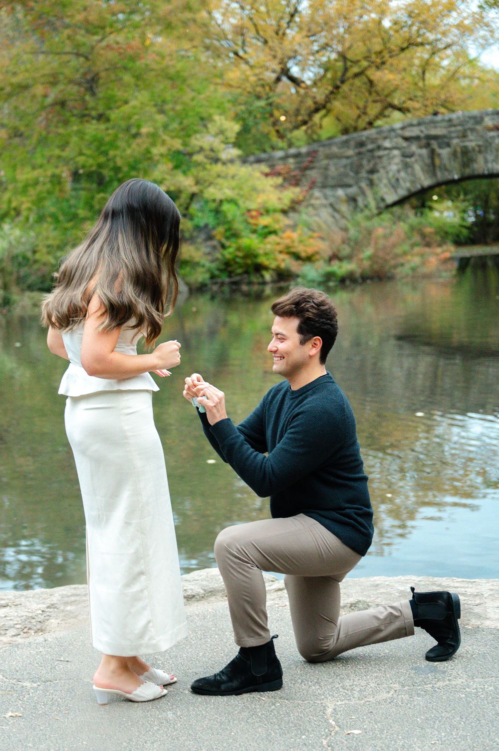 Smiling man on one knee presenting engagement ring to woman in white dress in Central Park.