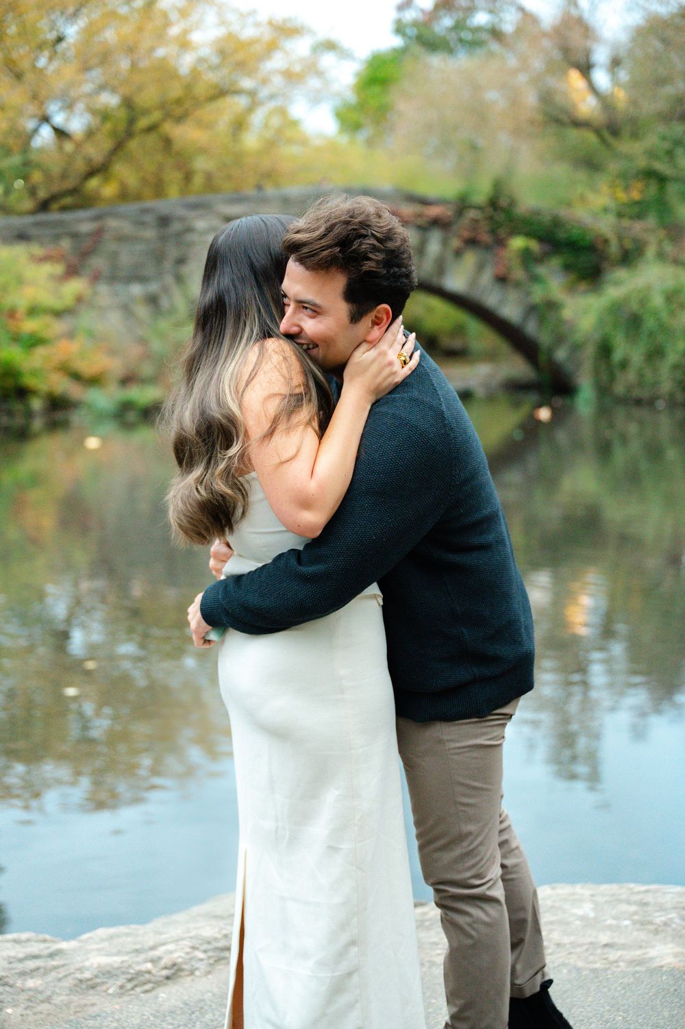 Newly engaged couple embracing and smiling by Central Park pond with stone arch bridge backdrop.