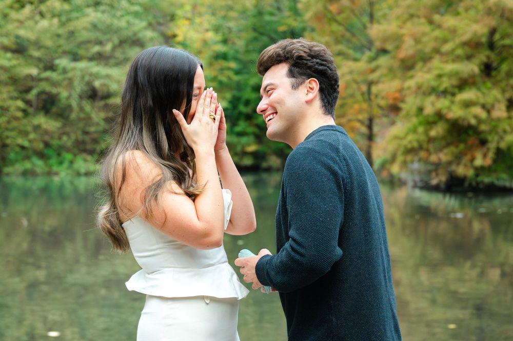 Couple sharing a joyful proposal moment outdoors near a lake surrounded by autumn foliage, woman covering her mouth in surprise.