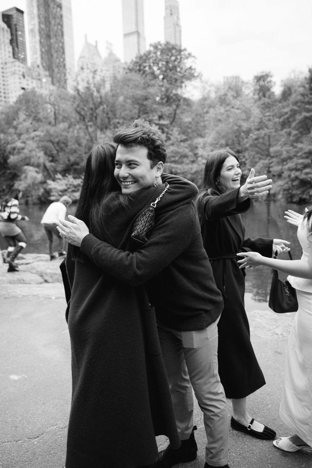 Black and white photo of joyful couple embracing after proposal in Central Park with friends celebrating nearby.