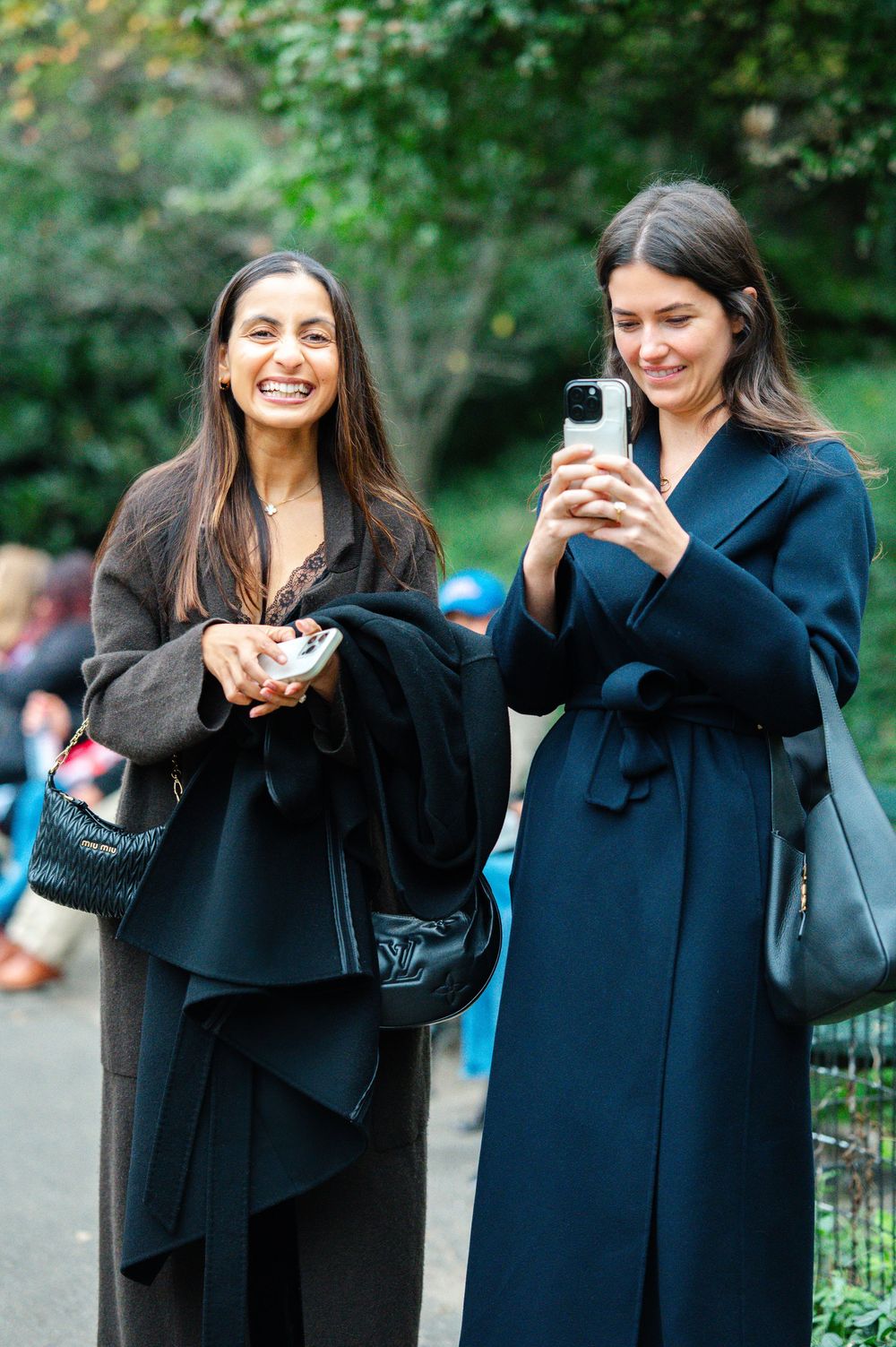 Two stylish women laughing outdoors, one taking photos on smartphone, wearing elegant dark coats in a park setting.