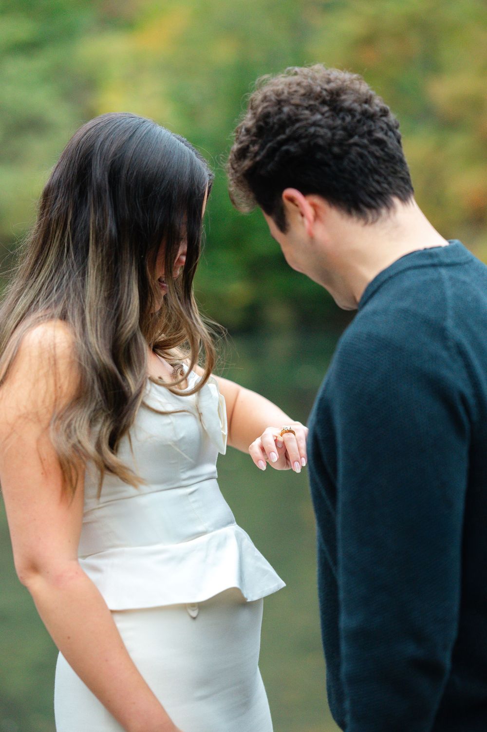 Couple admiring an engagement ring outdoors near a serene lake in autumn.