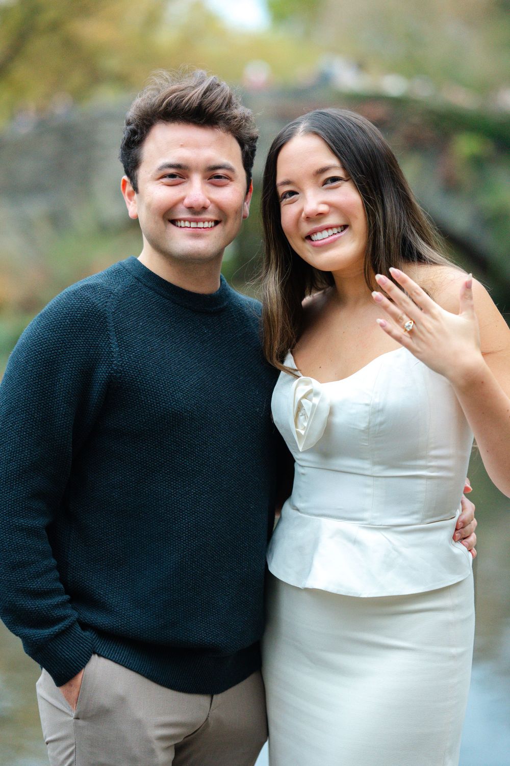 Smiling engaged couple posing by Central Park pond, woman proudly displaying her engagement ring.