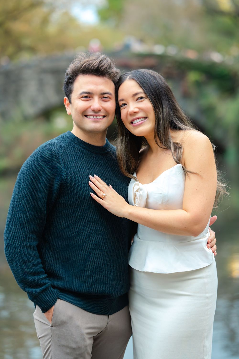 Happy couple posing for engagement photos near a bridge in Central Park, woman wearing white dress with ring visible.